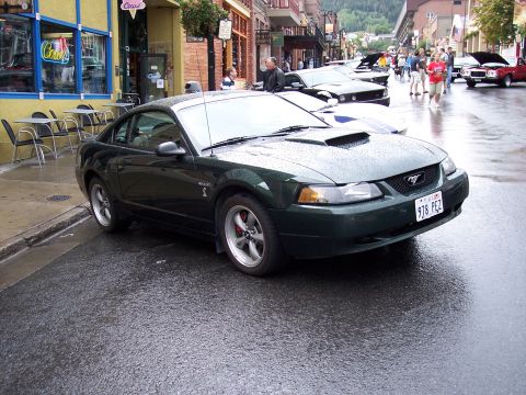 1972 Ford Ranchero - A dark green Ford Mustang is parked on a wet street in a town with shops and mountains in the background.