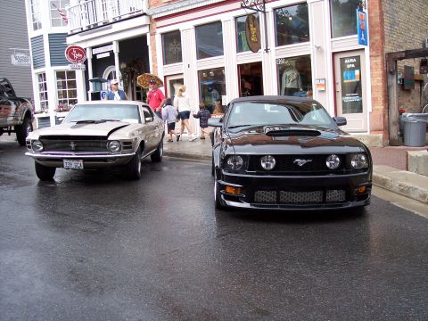 1972 Ford Ranchero - Two Ford Mustangs are parked on a wet street in front of shops, with people walking by.