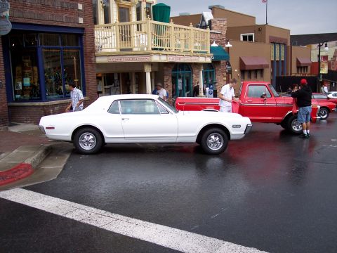1972 Ford Ranchero - Two classic vehicles, a white Mercury Cougar and a red Ford pickup, are parked on a wet street in front of shops.