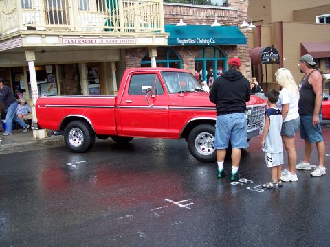 1972 Ford Ranchero - A red pickup truck is parked on a wet street in front of shops, with people observing.
