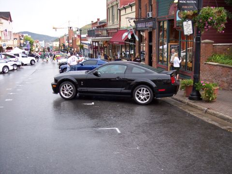 1972 Ford Ranchero - Black 2005 Ford Mustang parked on a wet street in a town with shops and other vehicles.