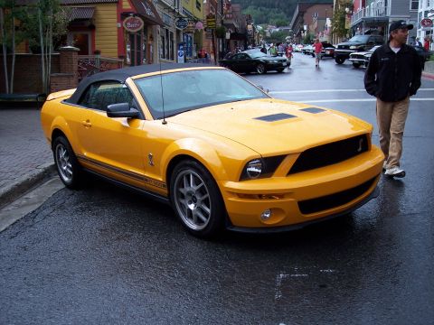 1972 Ford Ranchero - A yellow convertible Mustang Shelby GT500 is parked on a wet street in a town with shops.