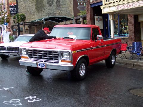 1972 Ford Ranchero - A red Ford pickup truck is parked on a wet street in front of shops, with a person cleaning its hood.