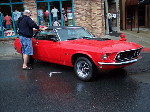 1972 Ford Ranchero - A red coupe with a black vinyl roof and hood scoop is parked on a wet street in front of a store.