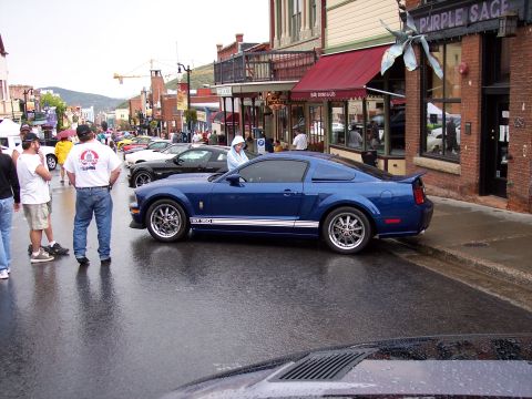 1972 Ford Ranchero - A blue Ford Mustang is parked on a wet street in front of shops.