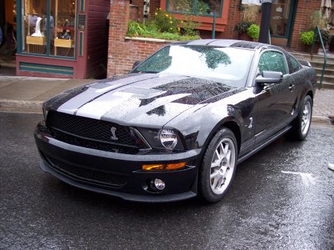 1972 Ford Ranchero - A black Ford Mustang Shelby GT500 with racing stripes is parked on a wet street in front of shops.