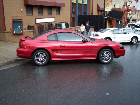 1972 Ford Ranchero - A red coupe with racing stripes is parked on a wet street next to other vehicles.