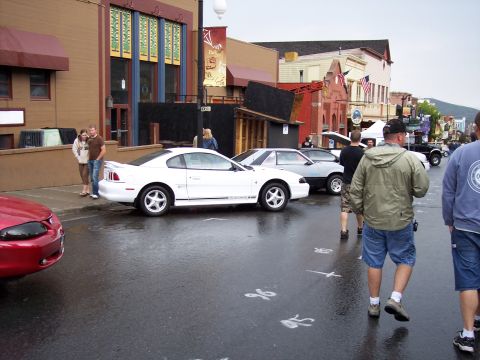 1972 Ford Ranchero - A white coupe with racing stripes is parked on a wet street in front of buildings.