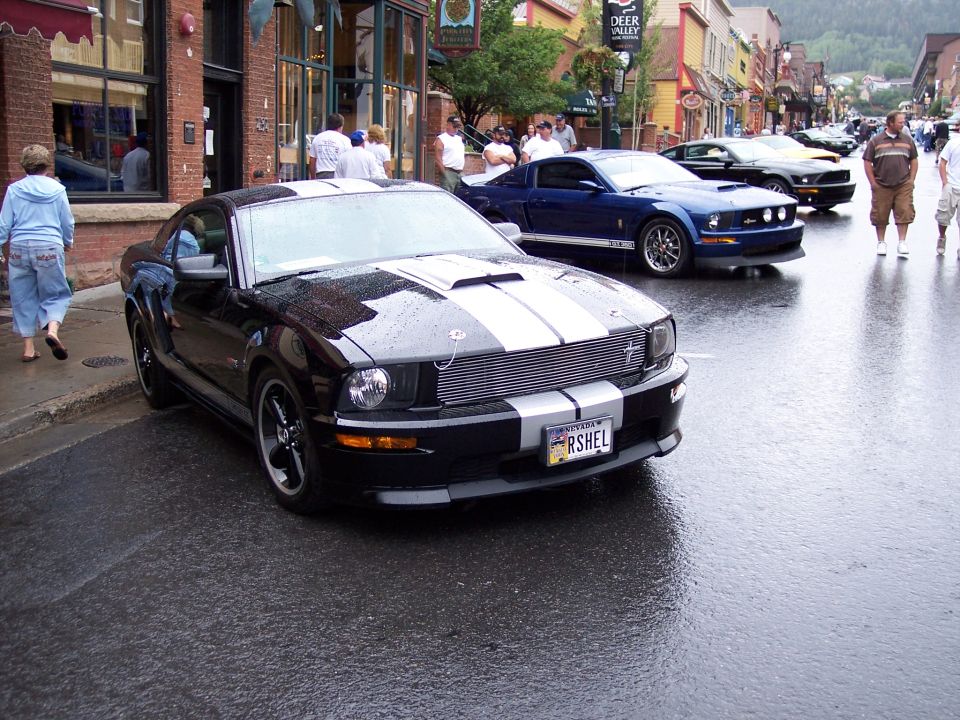 Black Mustang coupe with silver stripes parked on wet street near a blue Mustang