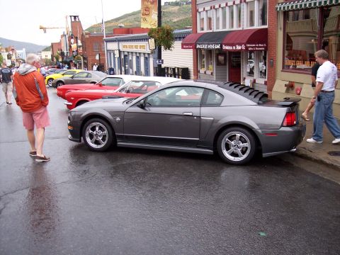 1972 Ford Ranchero - A row of cars are parked on a wet street in front of shops, with people walking on the sidewalk.