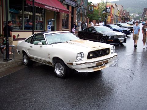 White Mustang II Cobra II coupe with gold stripes on a wet downtown street.