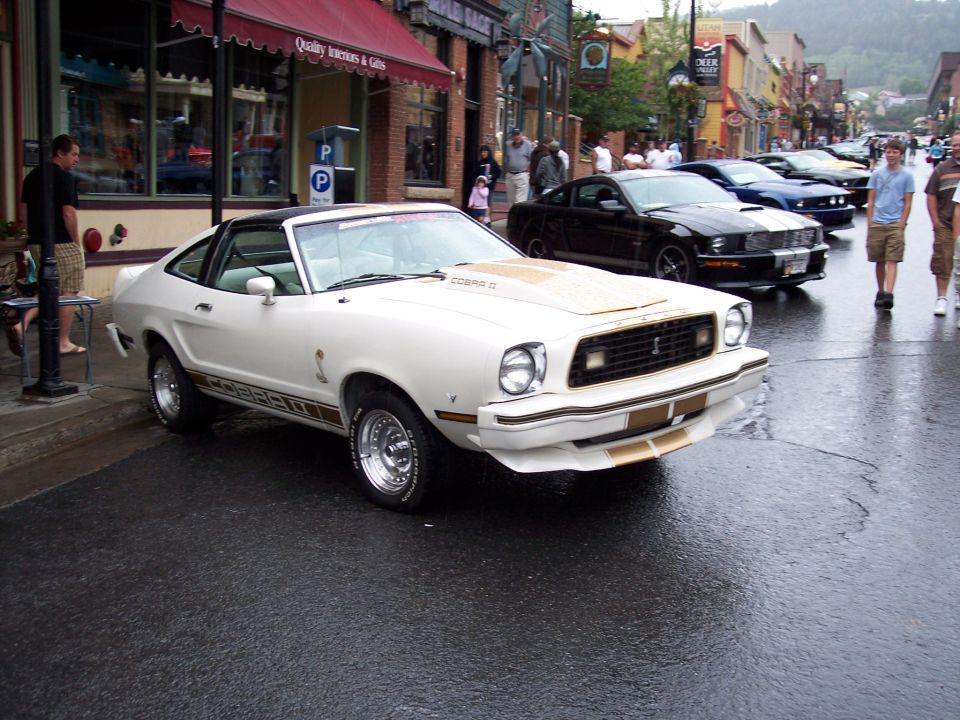 White Mustang II Cobra II coupe with gold stripes on a wet downtown street.
