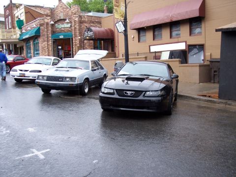 Black Ford Mustang coupe parked on wet street with older Mustang and white car behind.