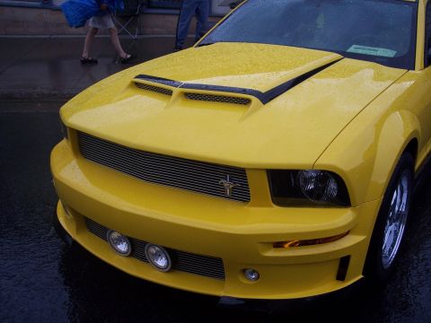 Rain-covered yellow Ford Mustang front three-quarter view with hood scoop and pony grille emblem