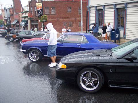 Blue classic Mustang coupe at a rainy street car show with black and red Mustangs nearby