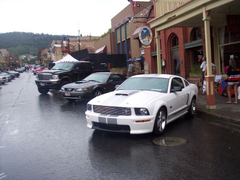 White Mustang coupe with gray stripes parked on wet downtown street at a car show