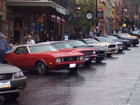 Row of parked Ford Mustangs, red convertible in front, on a wet downtown street