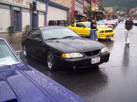 Black Mustang coupe with chrome wheels parked on wet street at a car show