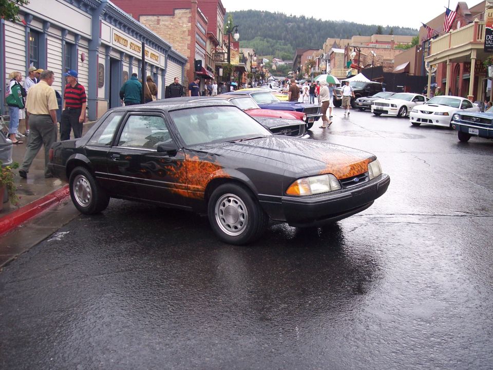 Black Fox-body Ford Mustang coupe with flame graphics on a wet street at a car show