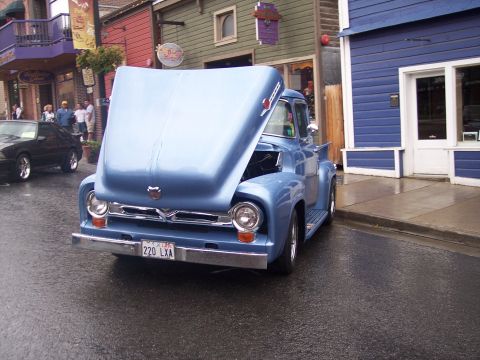 Light-blue 1950s Ford F-100 pickup with hood tilted open on a wet main street lined with shops