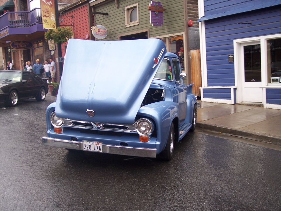 Light-blue 1950s Ford F-100 pickup with hood tilted open on a wet main street lined with shops