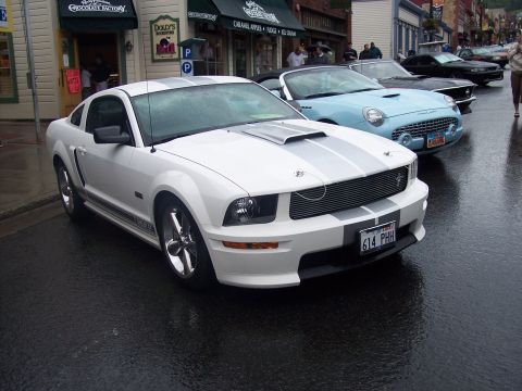 White Shelby GT Mustang with silver stripes on wet street, classic cars behind