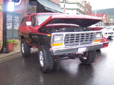 Lifted red and black Ford Bronco SUV with open hood on chrome wheels, parked on a wet street.