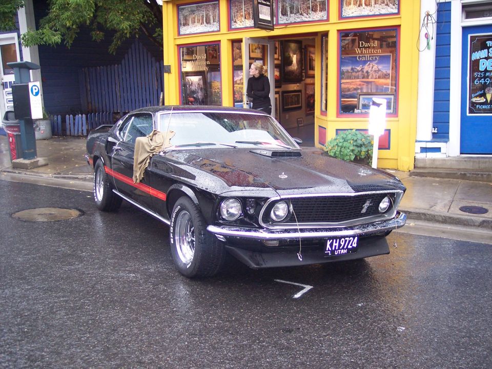 Black Ford Mustang fastback with red stripe parked wet on street by yellow art gallery
