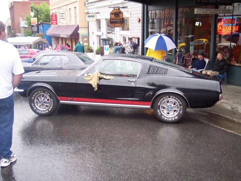 Black Mustang GT fastback in profile on wet street with chrome wheels and white-letter tires