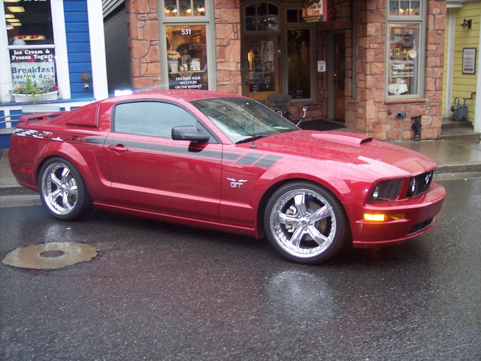 Dark red Ford Mustang GT coupe with black side stripes and chrome wheels parked on a wet street