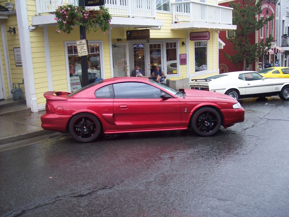 Red Mustang coupe in side profile on wet street with black wheels and rear spoiler