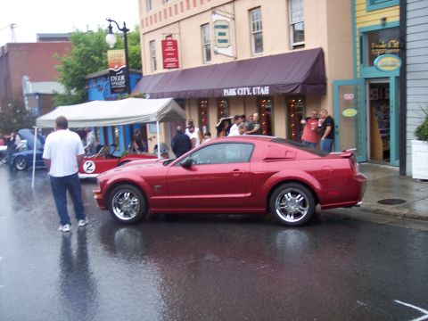 Red Ford Mustang coupe with chrome wheels parked on wet street at car show
