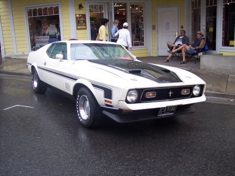 White Ford Mustang Mach 1 fastback with black hood, 3/4 front view on a wet street.