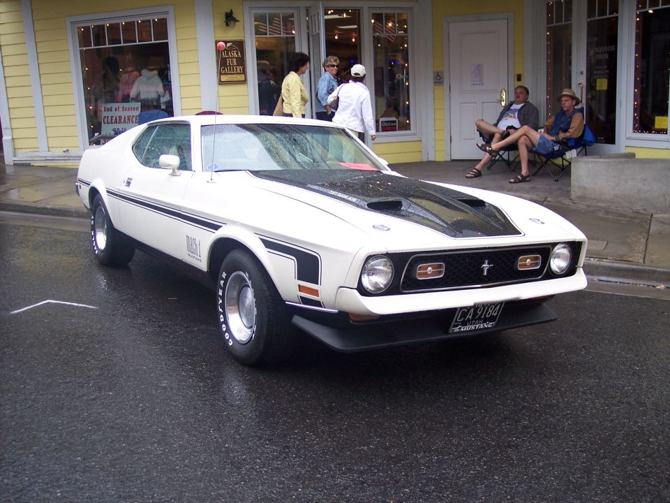 White Ford Mustang Mach 1 fastback with black hood, 3/4 front view on a wet street.