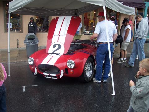 Red Cobra-style roadster with white stripes and number 2, hood up, at outdoor car show