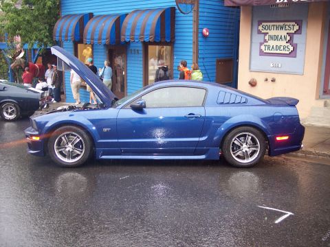 Blue GT coupe in profile with open hood on wet street at a car show.