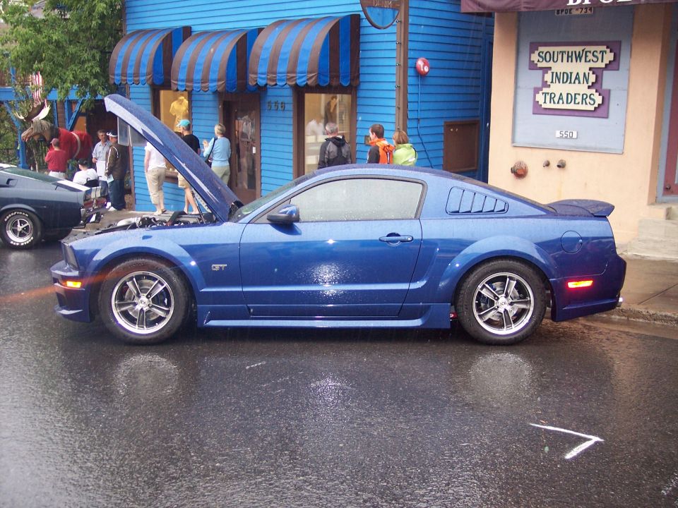 Blue GT coupe in profile with open hood on wet street at a car show.