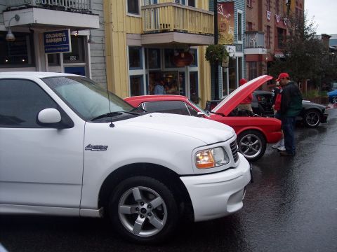 White Ford Lightning pickup at a wet street car show with a red truck and shops behind