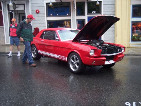Red coupe with white stripes and open hood on wet street, two men nearby