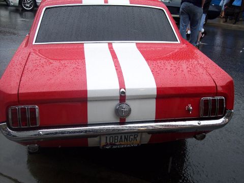 Rear of red Mustang with white stripes and '10BANGR' plate on wet pavement.