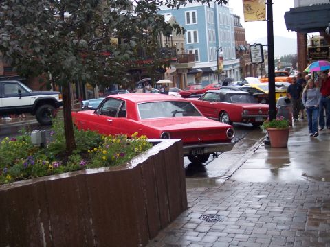Red coupe parked rear 3/4 view on a wet street with classic cars and umbrellas behind