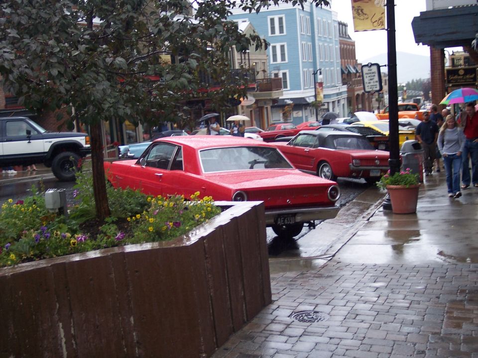 Red coupe parked rear 3/4 view on a wet street with classic cars and umbrellas behind