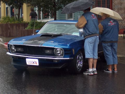 Blue Mustang coupe with black stripes and 'SIDEWDR' plate in the rain, two people with umbrellas beside it