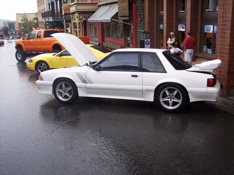 White Fox-body Mustang coupe with open hood and rear wing on wet street