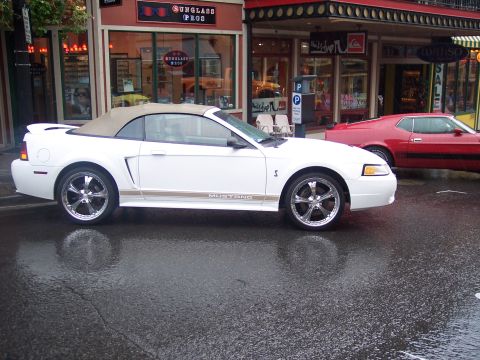 White Mustang convertible in profile on wet street with red classic car parked behind.