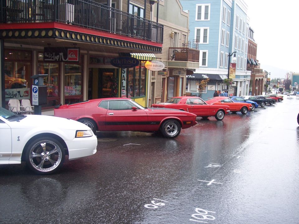 Row of Ford Mustangs from several generations parked on a wet downtown street in the rain