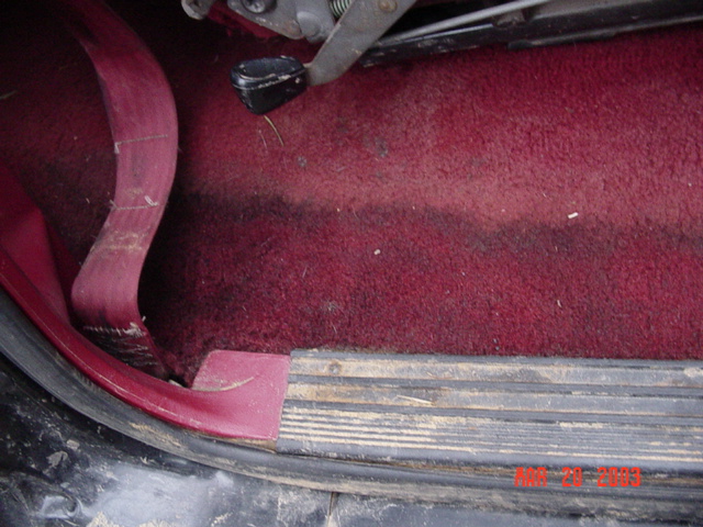 Maroon carpeted floorboard and door sill of a vehicle with hood-release handle visible