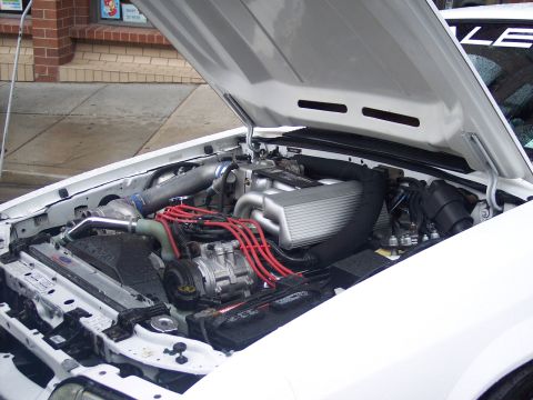 White car engine bay with red plug wires, aluminum intake and polished piping, hood up