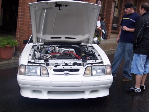 White Fox-body Mustang with open hood showing turbo engine, men standing beside it