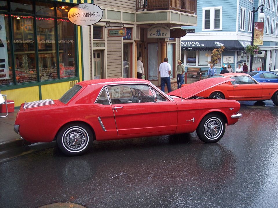 Red classic Mustang coupe, side view, on wet street with shops behind
